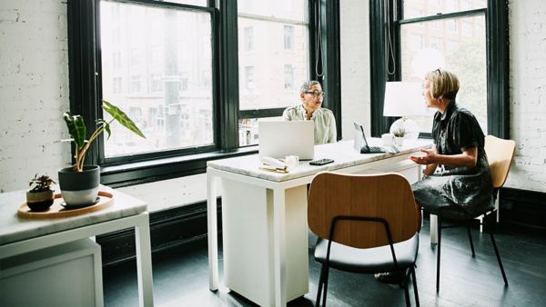A woman sitting at her computer talking to another woman.