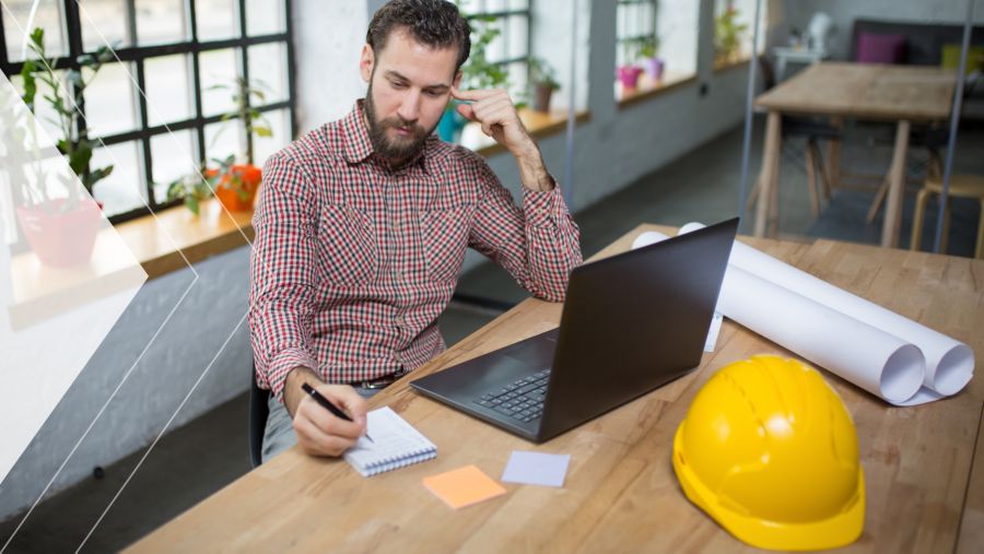A construction worker sits at his desk and does calculations while writing. 