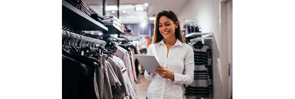 A retail worker looks at a tablet while standing next to a rack of retail clothing