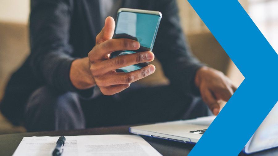 Man holding cellphone next to open laptop and documents on table 