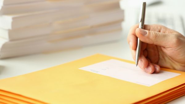 A hand holding a pen is shown above a stack of large yellow envelopes