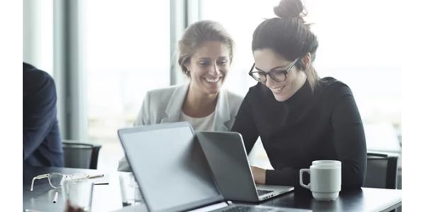 Two women sit at a conference table looking at a laptop discussing campaign solutions.