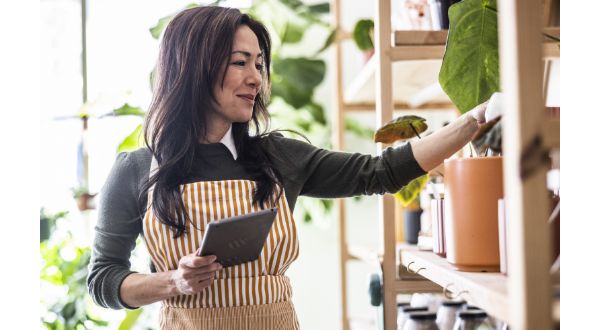 A small business owner tends to items on a shelf while holding a tablet.